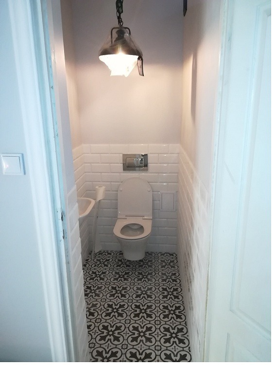 Wąska toaleta po remoncie: białe, cegiełkowe płytki na ścianach do połowy wysokości, wzorzysta czarno-biała podłoga, wisząca lampa w stylu industrialnym.