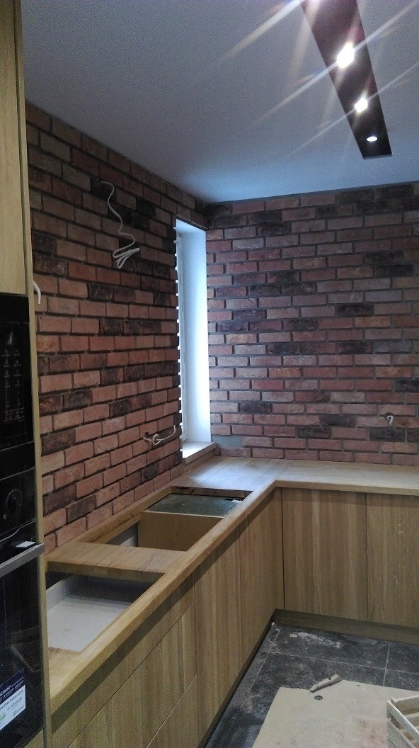 Kuchnia w trakcie wykańczania z ceglaną ścianą i drewnianymi szafkami, widoczne przewody elektryczne na ścianie.