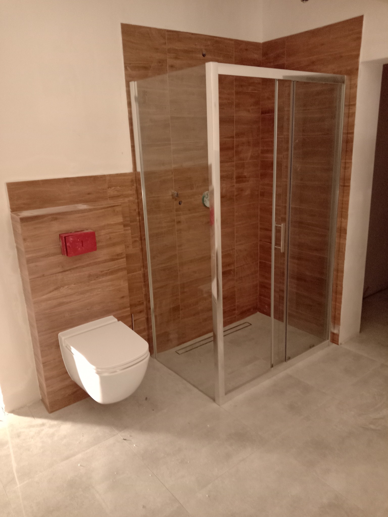 Nowoczesna łazienka w trakcie wykańczania: kabina prysznicowa z przesuwnymi drzwiami, podwieszana toaleta z czerwoną armaturą, płytki imitujące drewno na ścianach i jasne płytki podłogowe.