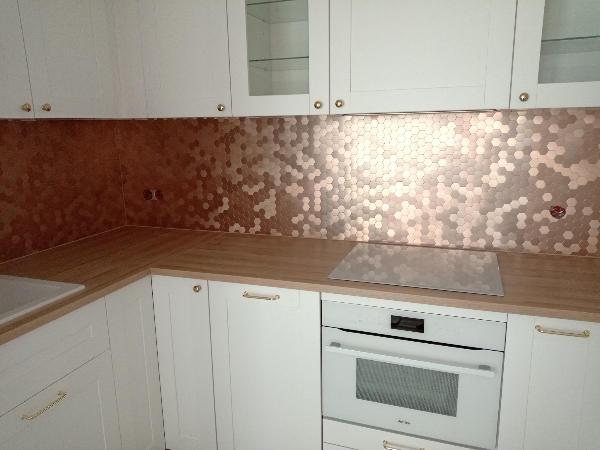 Kuchnia z białymi szafkami, drewnianym blatem i miedzianą mozaiką heksagonalną na ścianie nad blatem, z widoczną płytą indukcyjną i piekarnikiem marki Amica.