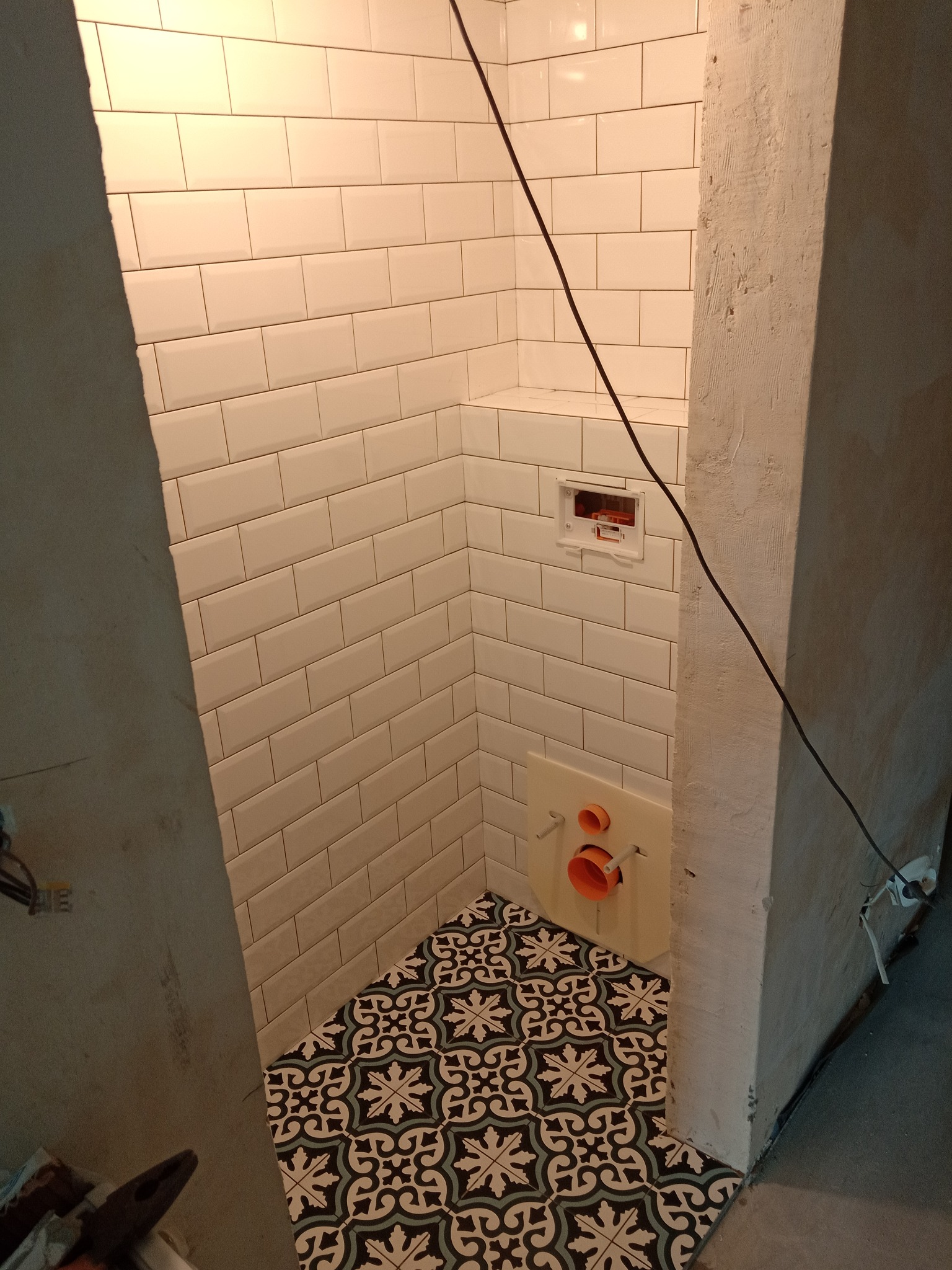 Fragment łazienki w trakcie remontu: białe, prostokątne płytki ścienne, wzorzysta podłoga w stylu retro, stelaż podtynkowy WC z pomarańczowymi rurami, surowy beton na ścianach.