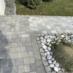 Porem - Szara kostka brukowa ułożona w geometryczny wzór, obrzeża z kamieni i roślinność ozdobna tworzą estetyczne przejście przy budynku.