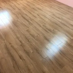 Gotowa podłoga z paneli laminowanych o ciepłym odcieniu drewna, ułożona w pokoju z różowymi ścianami, widoczne odbicie światła na powierzchni.