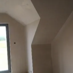 Pokój na poddaszu po nałożeniu tynku maszynowego, widoczne nierówności i surowy beton na podłodze.