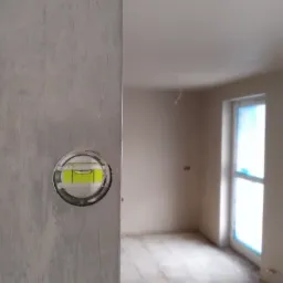 Poziomica przyłożona do ściany w surowym stanie, w tle widoczne okno zabezpieczone folią i fragment pomieszczenia w trakcie remontu.