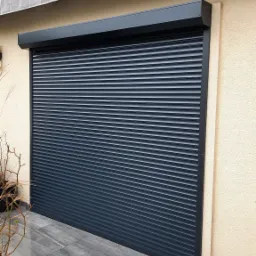 Zasunięta, grafitowa roleta na elewacji budynku, widok z zewnątrz na marmurowym tarasie, z donicą z rośliną po lewej stronie.