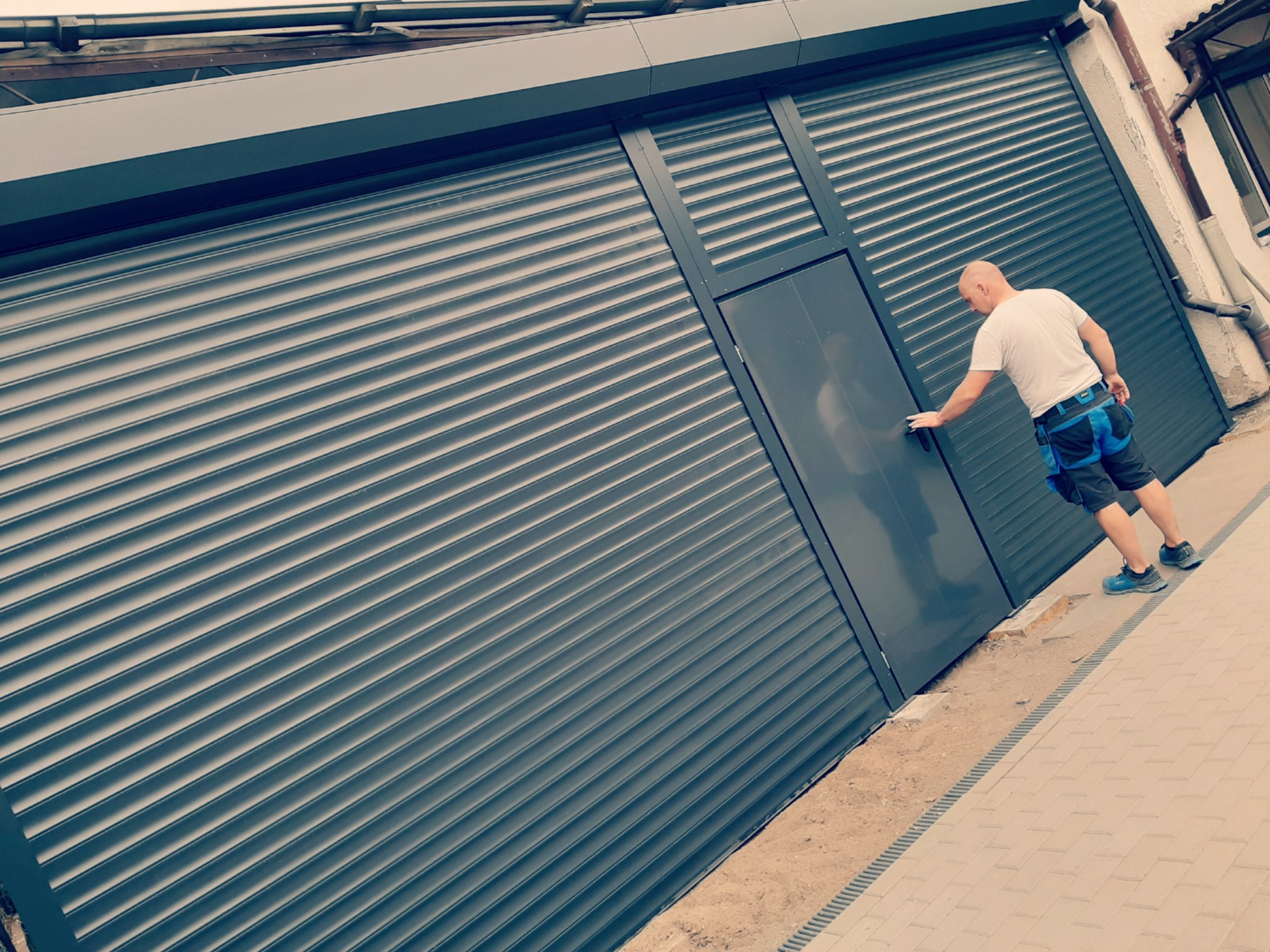 Mężczyzna w roboczym ubraniu dotyka ciemnoszarej bramy rolowanej z wbudowanymi drzwiami, widoczne elementy konstrukcyjne i fragment chodnika