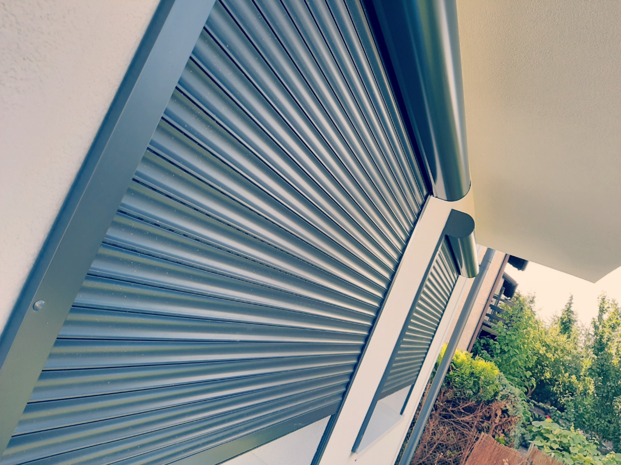 Zamknięta, szara roleta zewnętrzna z widocznym mechanizmem zwijania, zamontowana na elewacji budynku z widokiem na zieleń ogrodu.