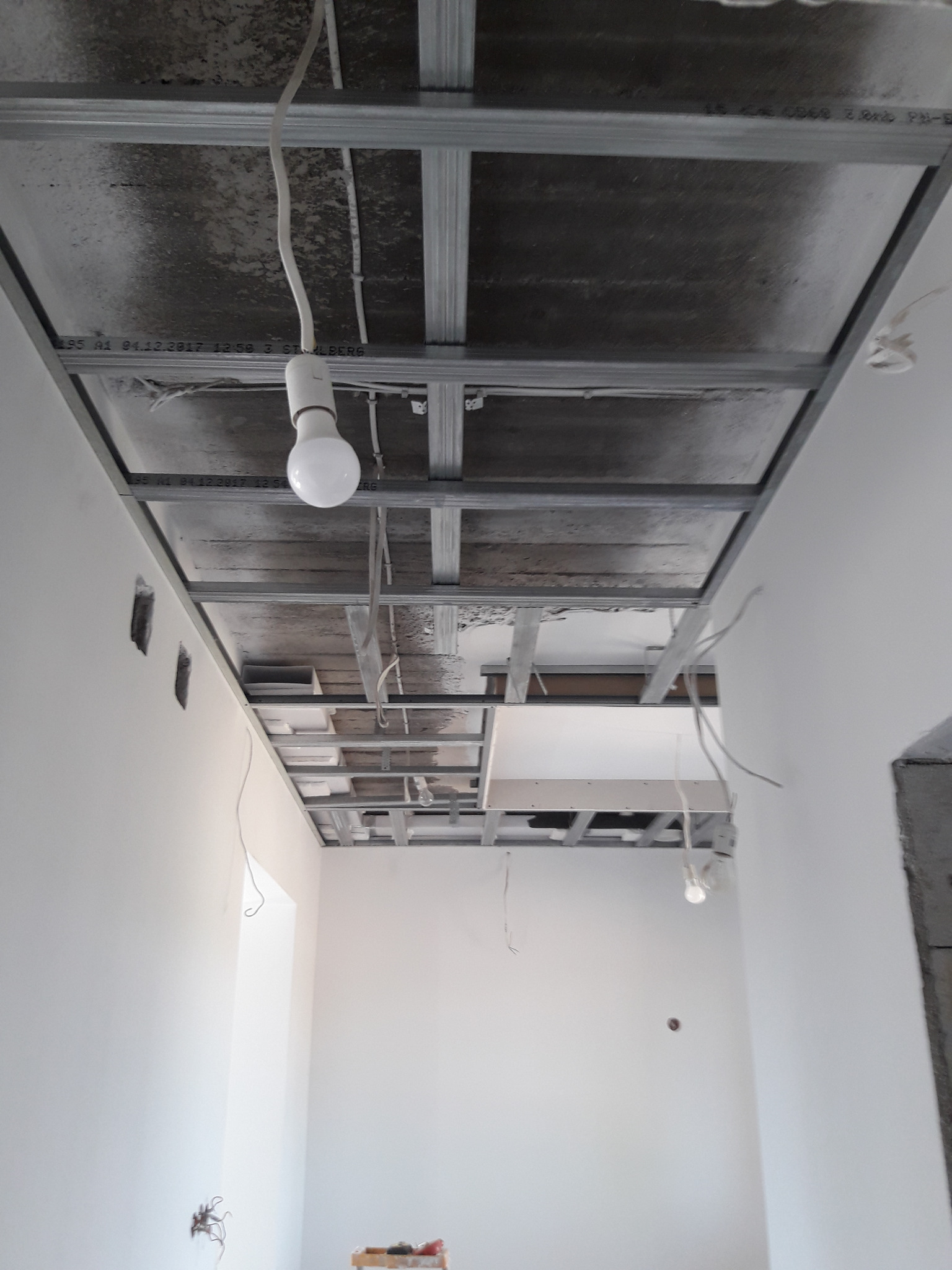 Sufit podwieszany ze stelażem z profili metalowych, widoczna instalacja elektryczna i wisząca żarówka, ściany w trakcie wykończenia, widoczne puszki elektryczne.