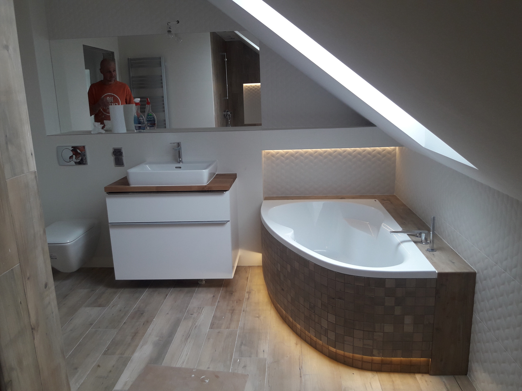 Nowoczesna łazienka na poddaszu z asymetryczną wanną obudowaną mozaiką, umywalką nablatową na szafce i drewnopodobną podłogą, widok z perspektywy wejścia.