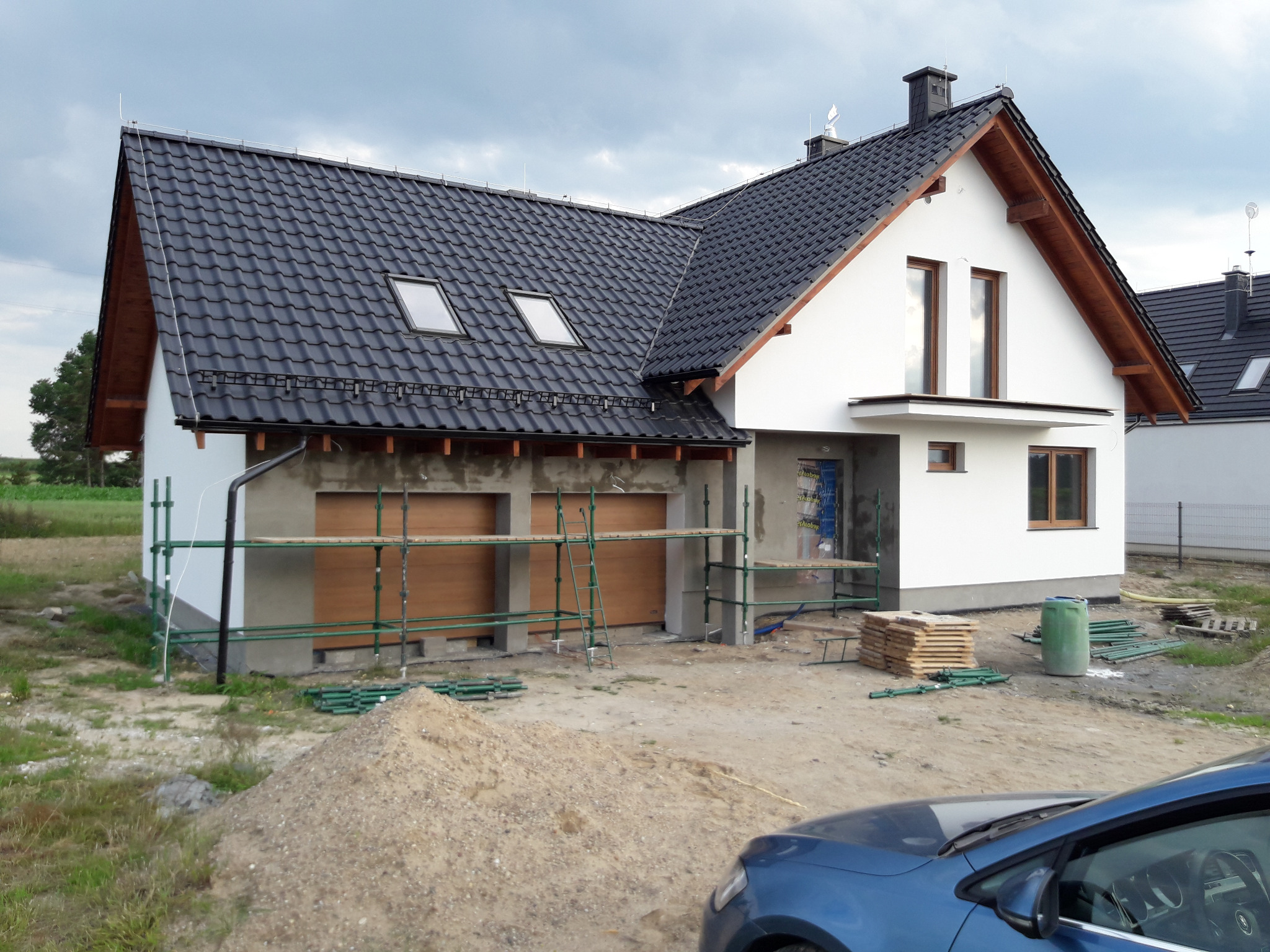 Nowy dom jednorodzinny w trakcie wykańczania elewacji, widoczne rusztowania, przygotowany teren i fragment auta na pierwszym planie.