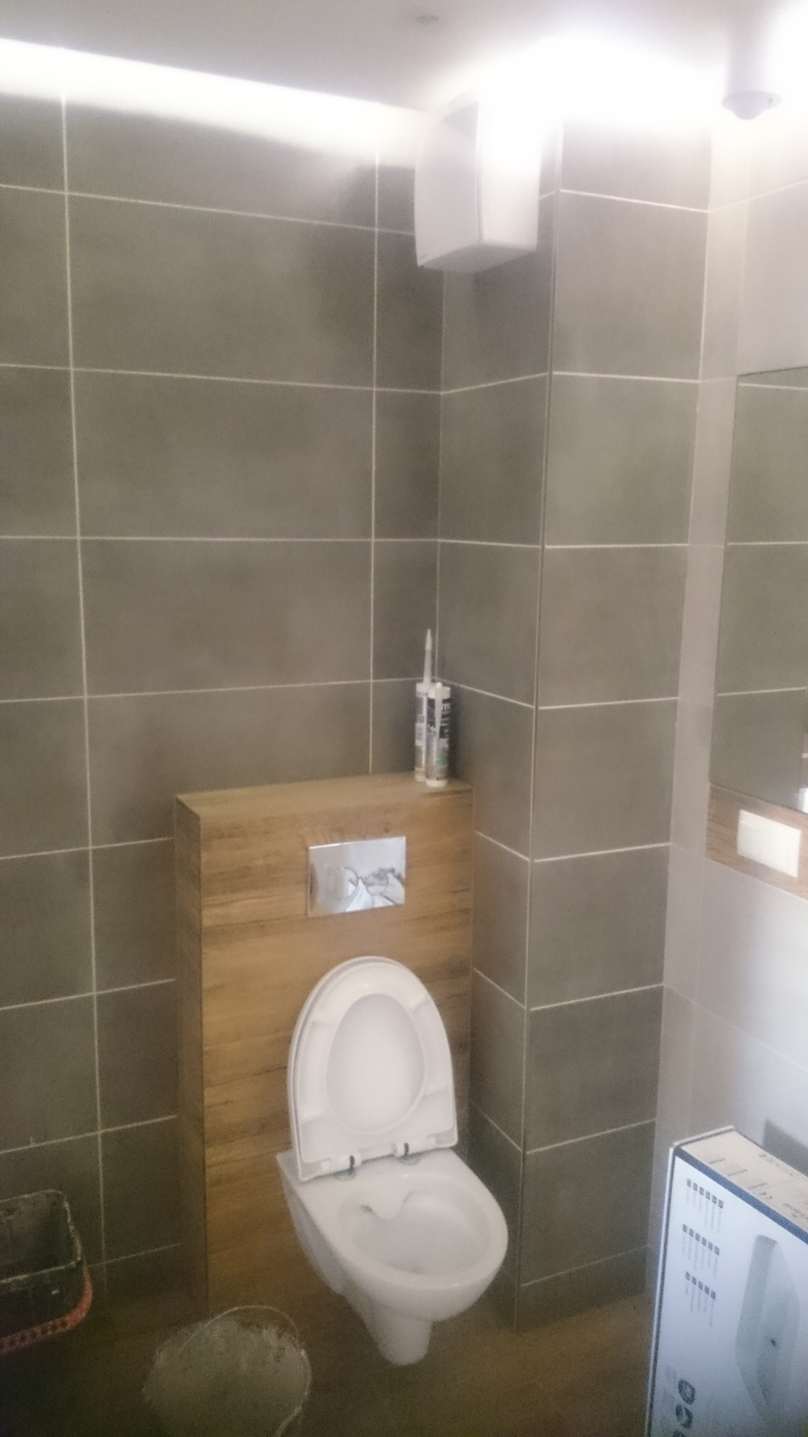 Wisząca toaleta z drewnianą obudową zbiornika, szare płytki na ścianach, tuby silikonu na obudowie, częściowo widoczny pojemnik i pudełko z armaturą w łazience.