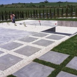 JULIA OGRODY - Nowoczesny ogród w trakcie realizacji: szare płyty chodnikowe ułożone w trawie, biały żwir, betonowe obrzeża, rząd tuj i pracownik z grabiami w tle.