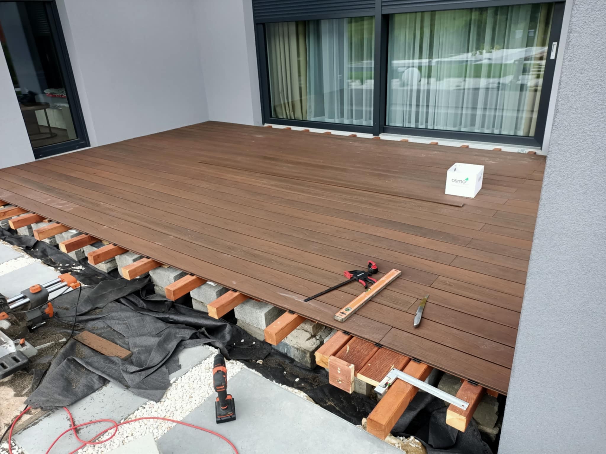 Montaż drewnianego tarasu: deski w odcieniach brązu układane na legarach, widoczne narzędzia (poziomica, wkrętarka), częściowo ułożona podkonstrukcja.