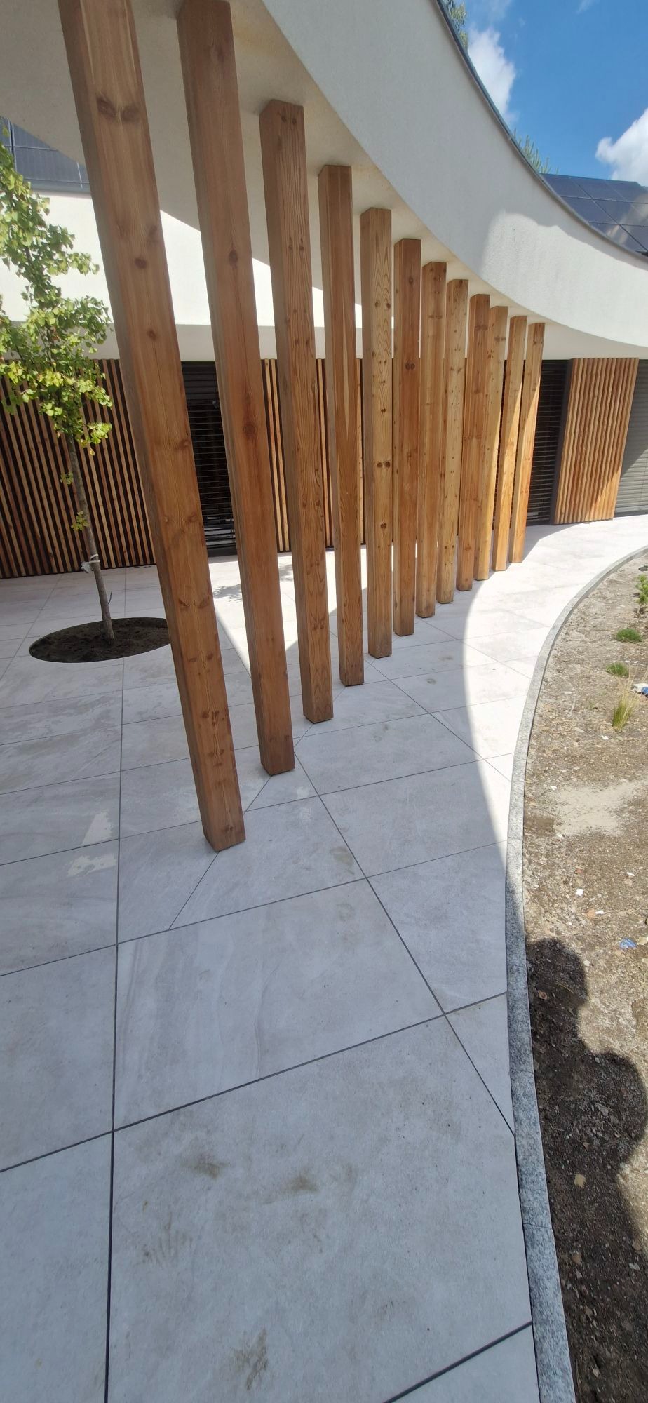 Nowoczesny ogród z drewnianą palisadą i jasną kostką. Widoczny fragment trawnika i drzewo. Architektura budynku z zaokrąglonymi elementami.