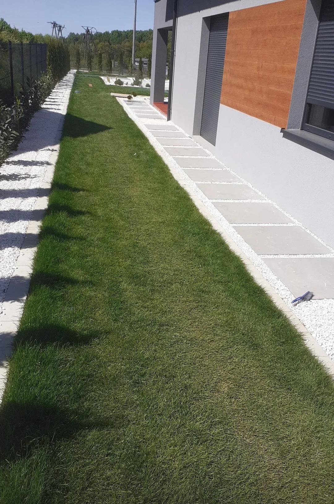 Ścieżka z płyt i białego żwiru prowadząca wzdłuż domu z drewnianą elewacją, obok zadbany trawnik. Widok na estetyczny ogród.