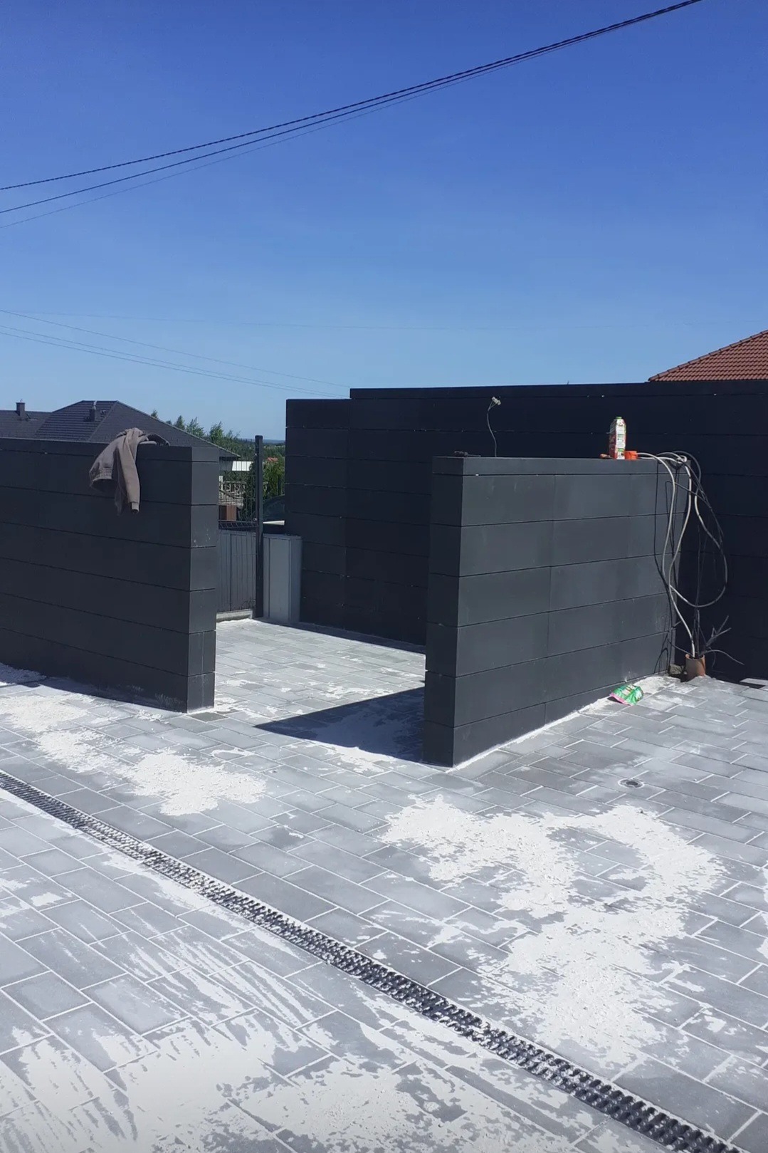 Nowoczesna, szara kostka brukowa z odpływem liniowym i ciemnymi panelami ogrodzeniowymi. Widoczne ślady po piasku lub cemencie. Słoneczny dzień.