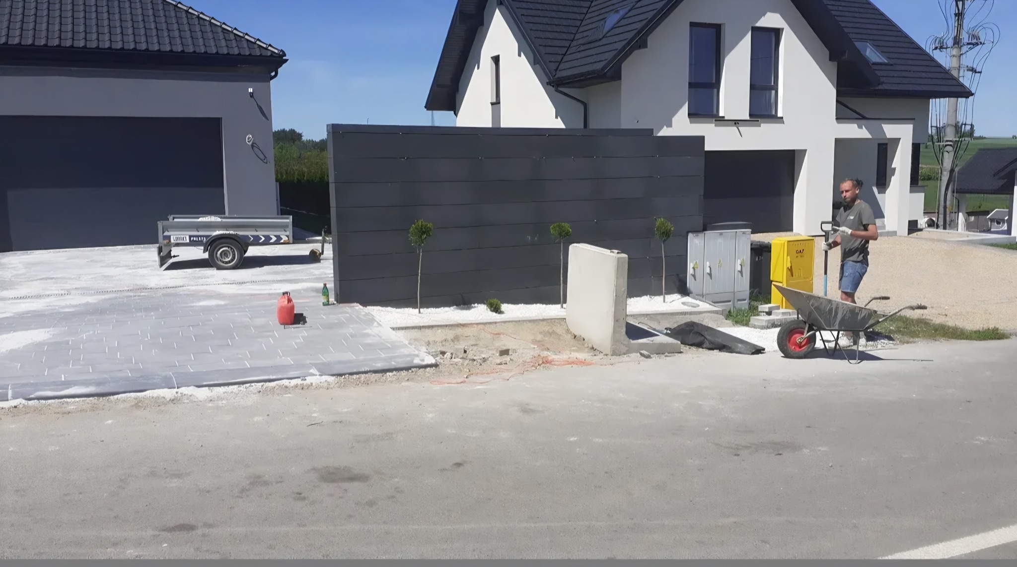 Układanie kostki brukowej na podjeździe, widoczny mur z nasadzeniami, pracownik z taczką i narzędziami, domy w tle. Prace w toku, dzień słoneczny.