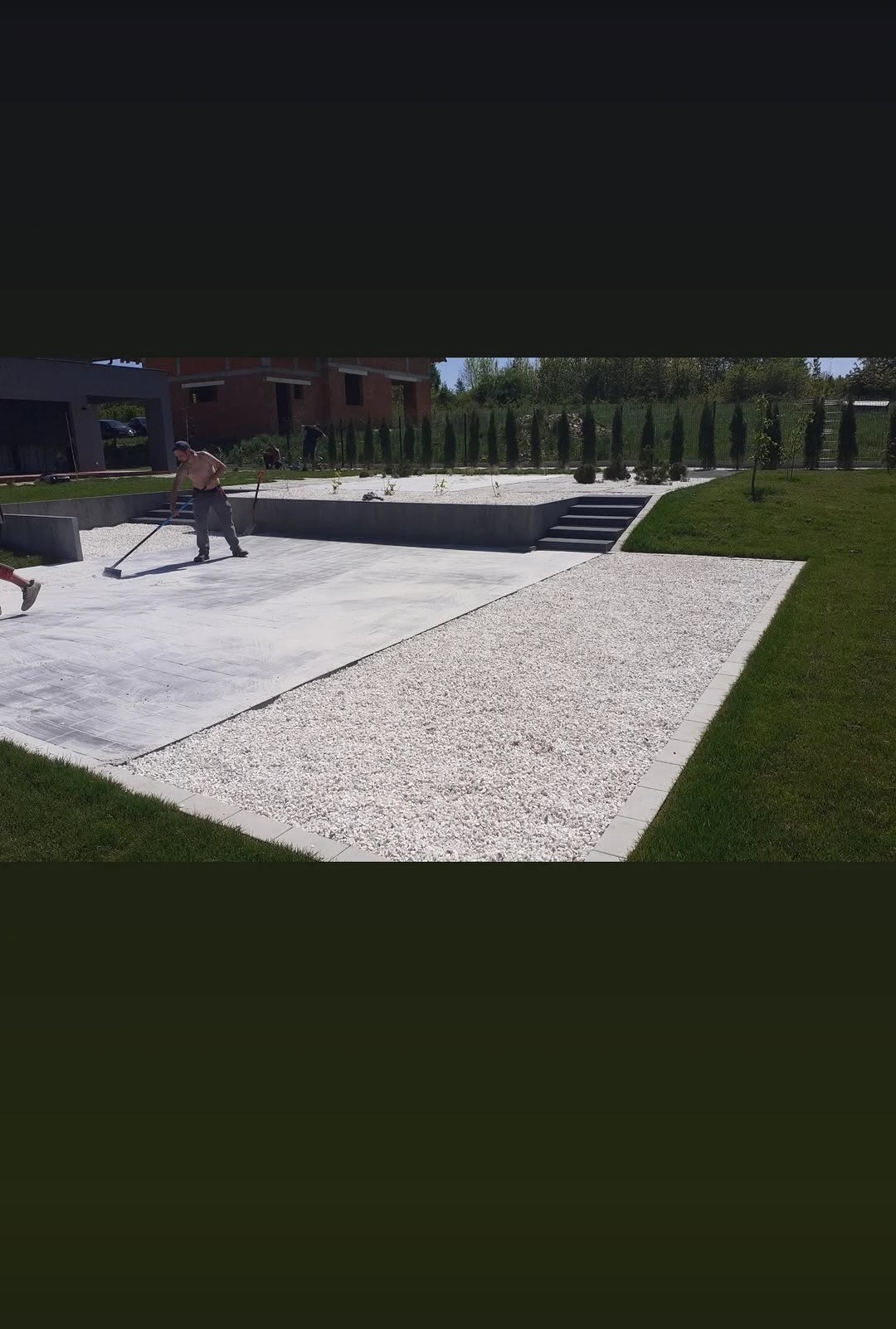 Nowoczesny ogród w trakcie realizacji: betonowa platforma, biały żwir, trawnik i nasadzenia. Pracownik wyrównuje nawierzchnię. Budynek w tle.