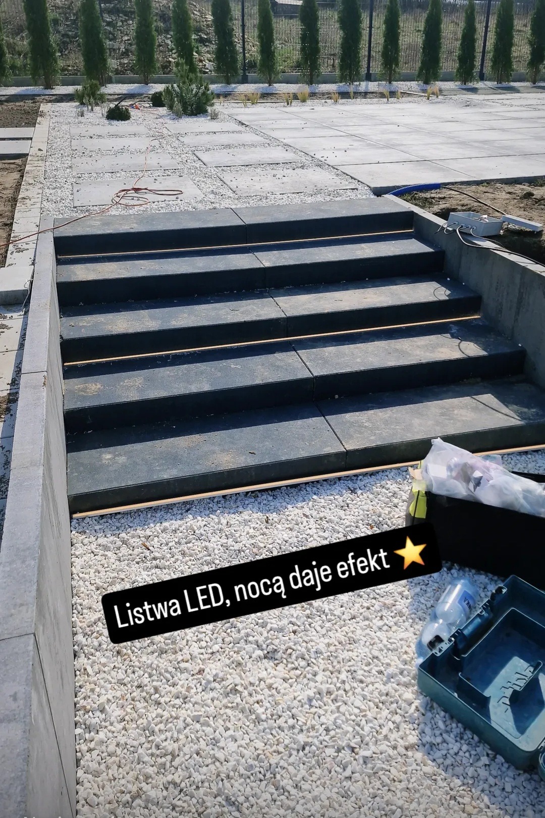 Schody z podświetleniem LED w ogrodzie, wykończone białym żwirem i betonowymi płytami. W tle rząd tuj i fragment trawnika.