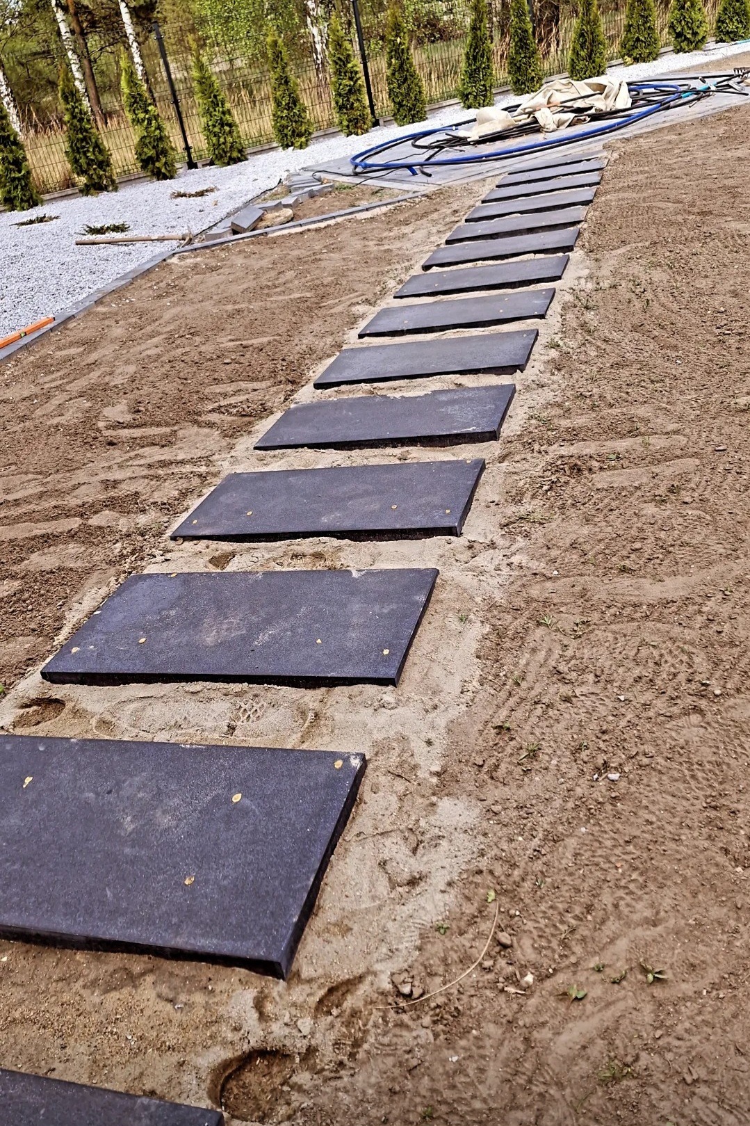 Prosta ścieżka z ciemnych płyt w ogrodzie, prowadząca przez piaskową nawierzchnię w kierunku zielonych drzew i krzewów. Widoczne ślady opon na ziemi.