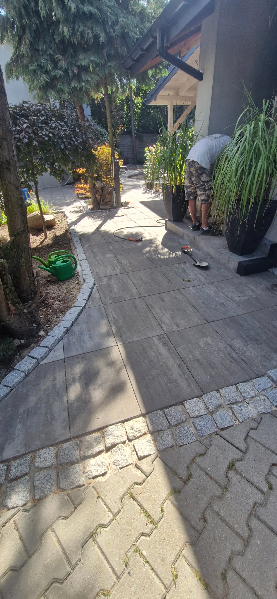 Wykończony chodnik z kostki brukowej obrzeżony kamieniami. Widoczne rośliny w donicach, narzędzia ogrodnicze i osoba doglądająca roślin. Elementy małej architektury.