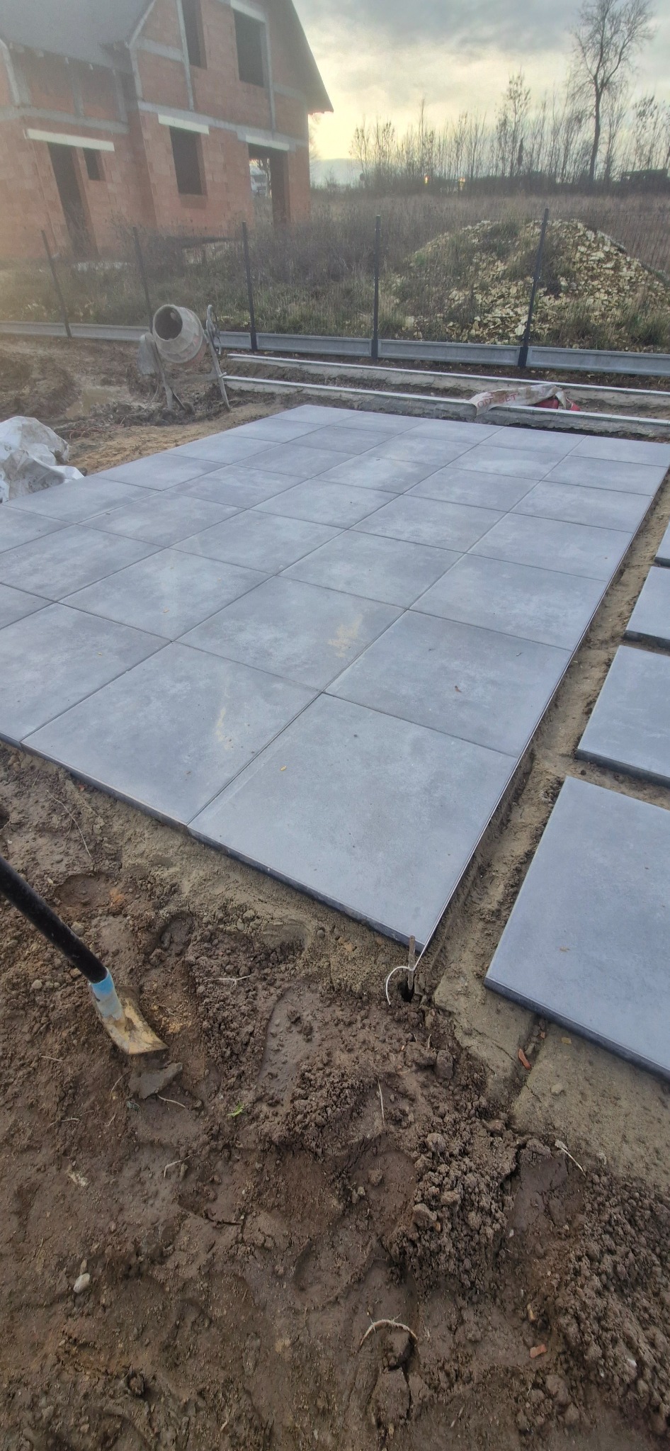 Ułożona, szara kostka brukowa na gruncie, widoczna łopata i fragmenty niezakończonej nawierzchni. W tle dom w stanie surowym i mieszarka do betonu.