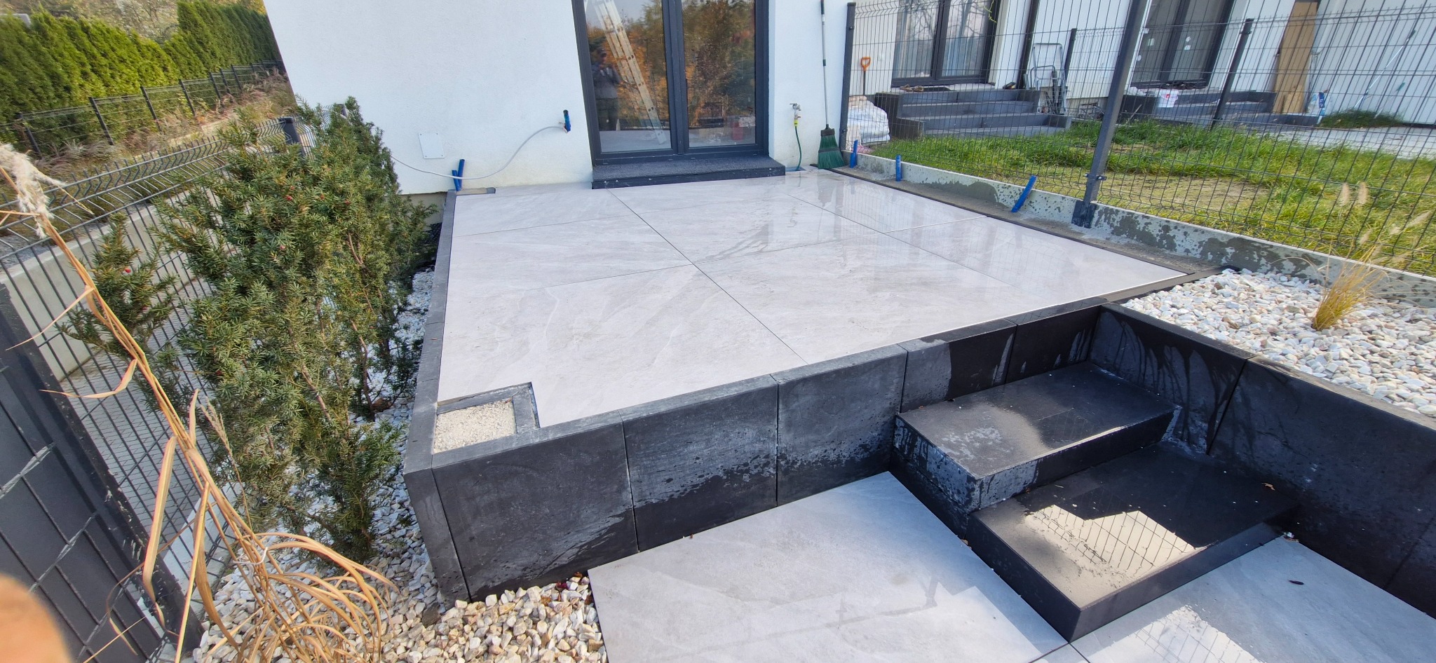 Wykończony taras z jasnych płyt, obrzeża z ciemnych bloczków, schody. Widoczne rośliny iglaste i ozdobne trawy. Nowoczesny design, schludne wykonanie.