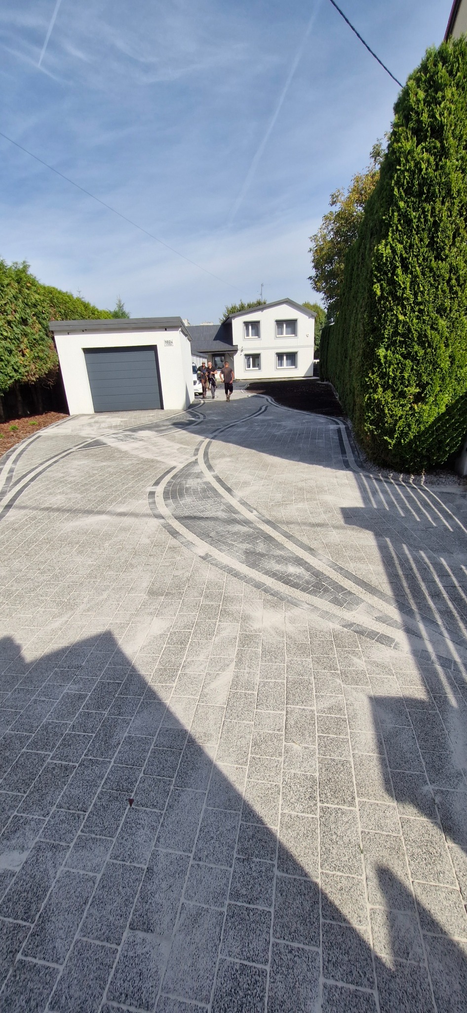 Ułożona kostka brukowa w odcieniach szarości tworząca wzór na podjeździe przed domem z garażem. Widoczne tuje i fragment nieba.
