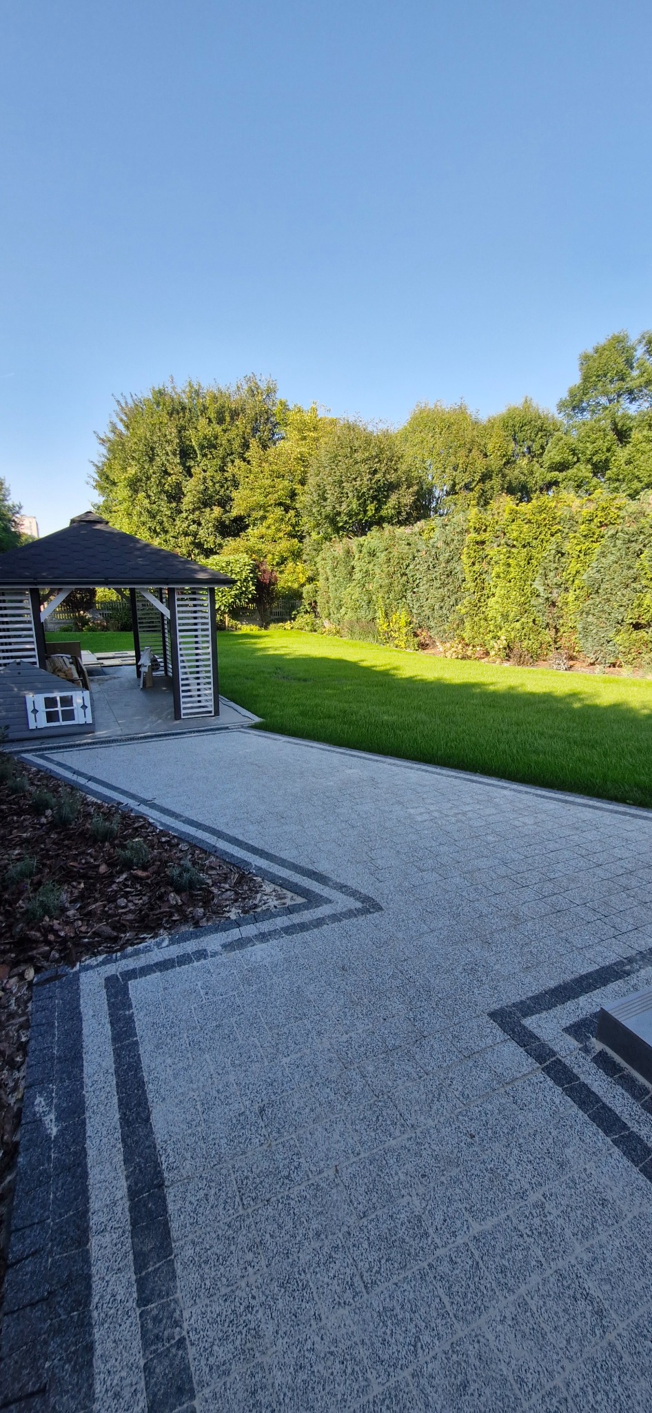 Ogród z altaną i ścieżką z kostki brukowej w geometryczny wzór, świeżo skoszony trawnik, drzewa i krzewy w tle. Słoneczny dzień.