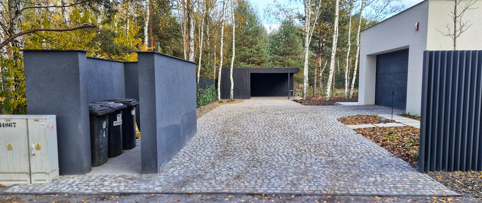 Ułożona kostka brukowa na podjeździe do nowoczesnego domu z garażem i wiatą, obok drzewa i zabudowa śmietnikowa w szarym kolorze.