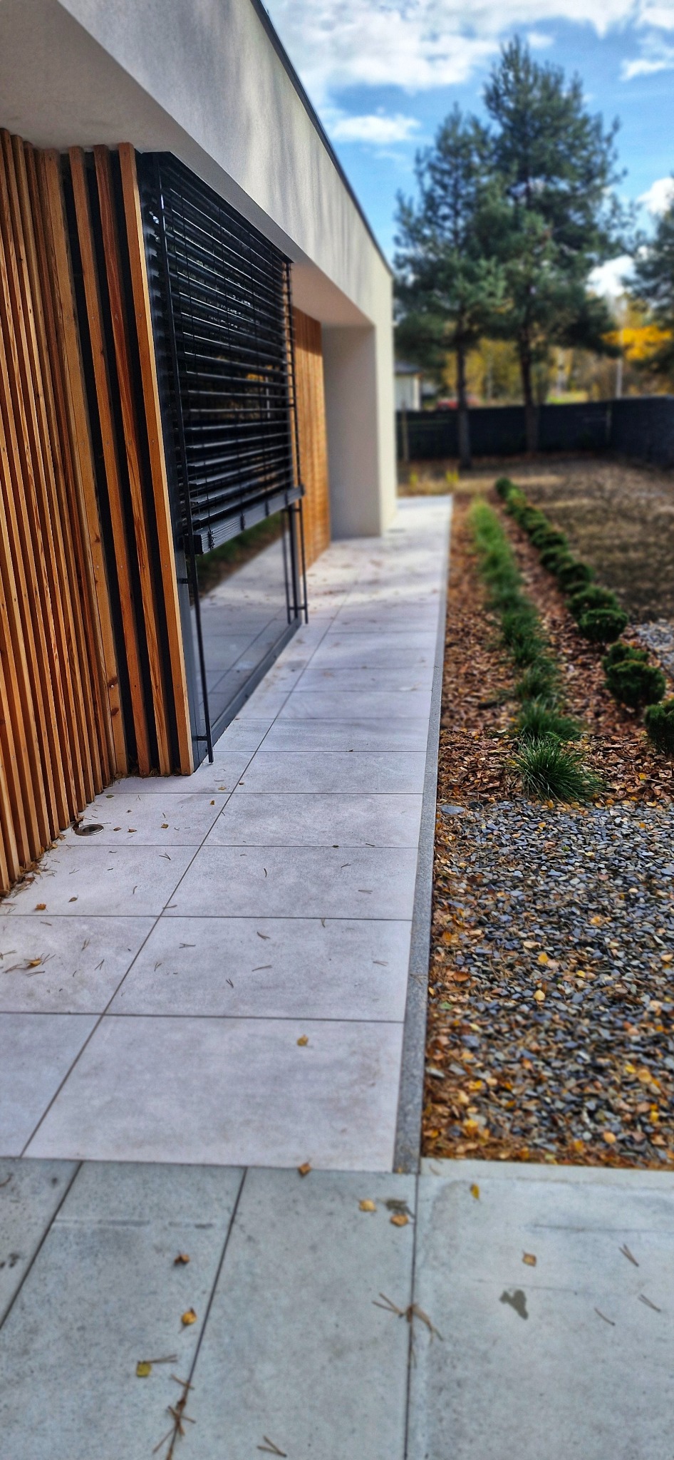 Nowoczesny ogród z szarym chodnikiem z płyt, obok rabata z kamykami i niskimi krzewami. Drewniana elewacja budynku w tle, jesienne drzewa.