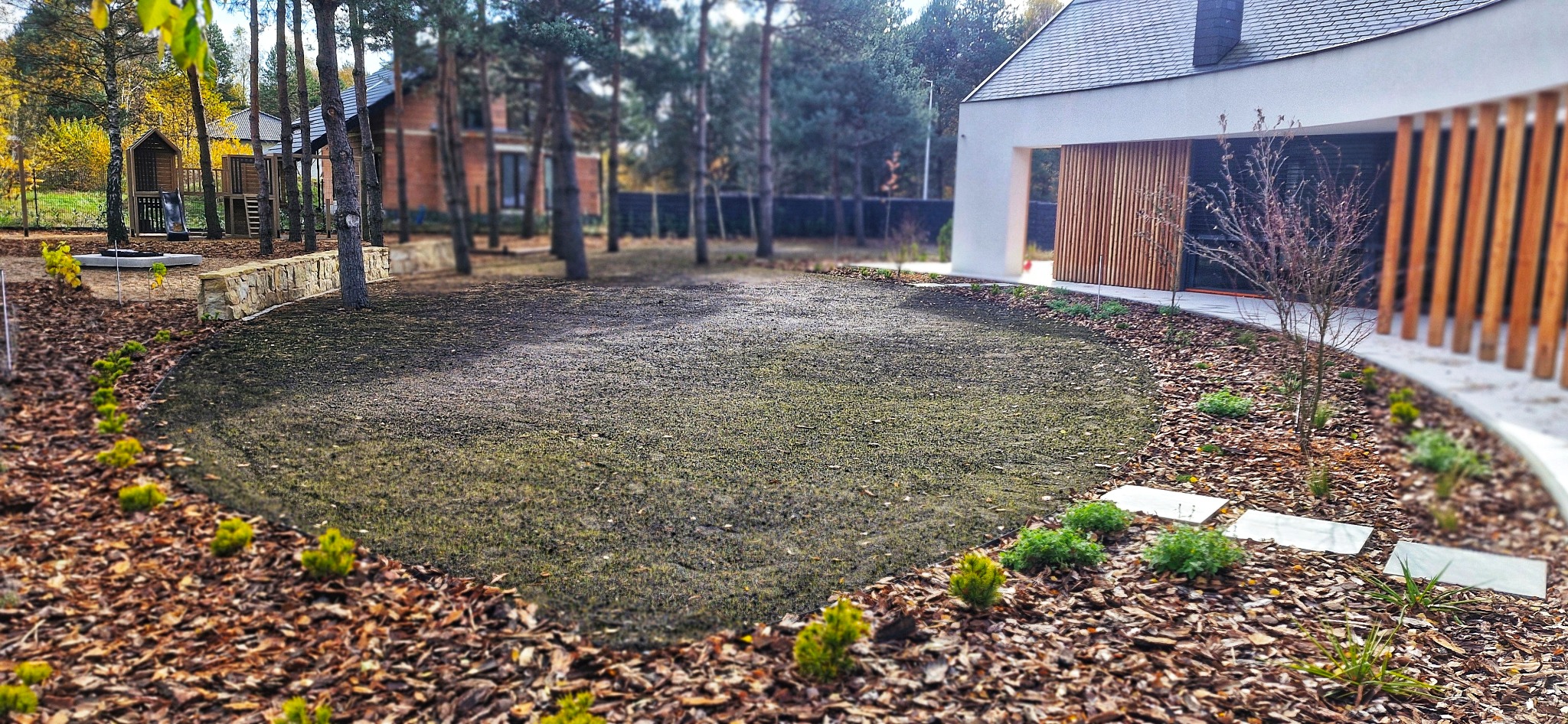 Świeżo założony trawnik z młodymi roślinami i korą drzewną, otoczony drzewami i nowoczesnym domem z drewnianą fasadą. Widok na plac zabaw w tle.