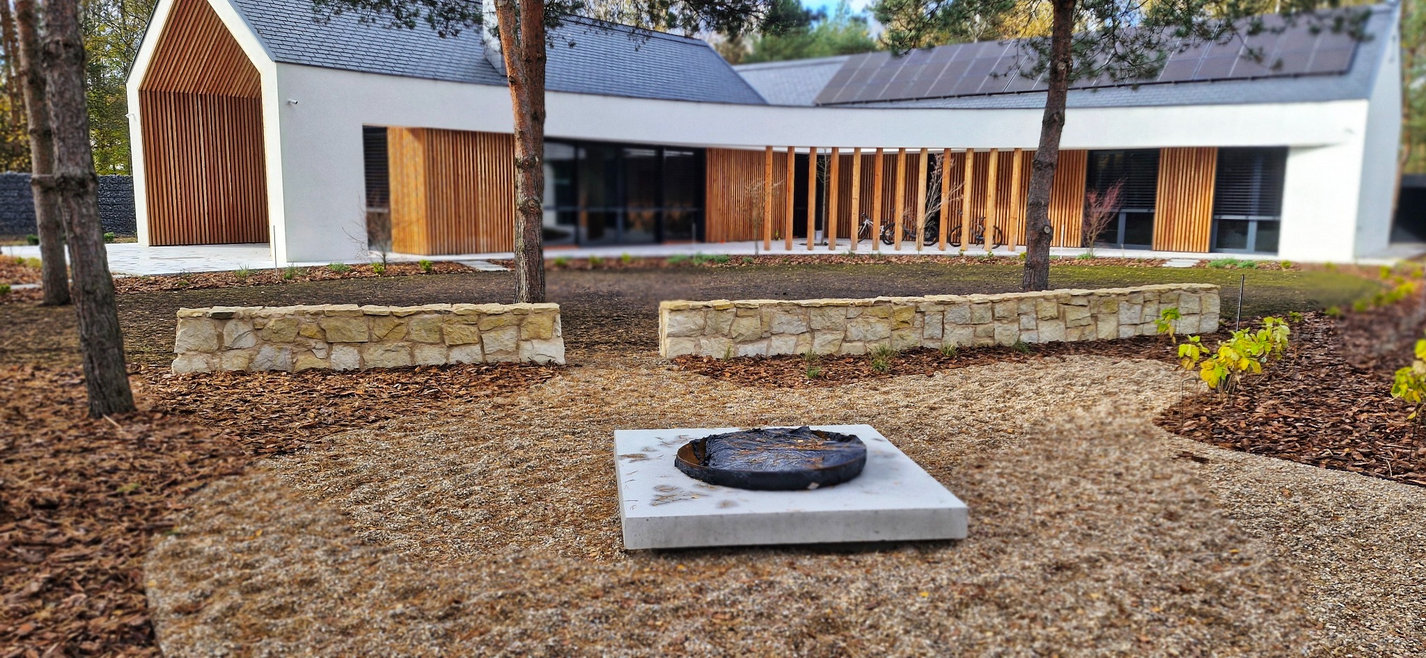 Nowoczesny ogród z minimalistycznym paleniskiem na tle domu z drewnianą elewacją i panelami słonecznymi. Kamienne murki oddzielają rabaty.