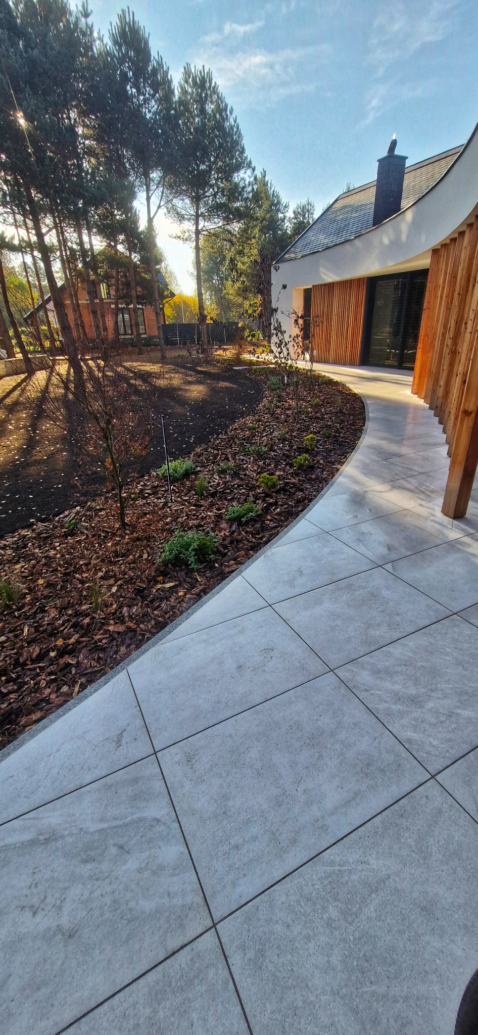 Nowoczesny ogród z jasnoszarą, kwadratową ścieżką z płytek, świeżo nasadzoną roślinnością i korą. W tle dom z drewnianą elewacją i wysokie drzewa.