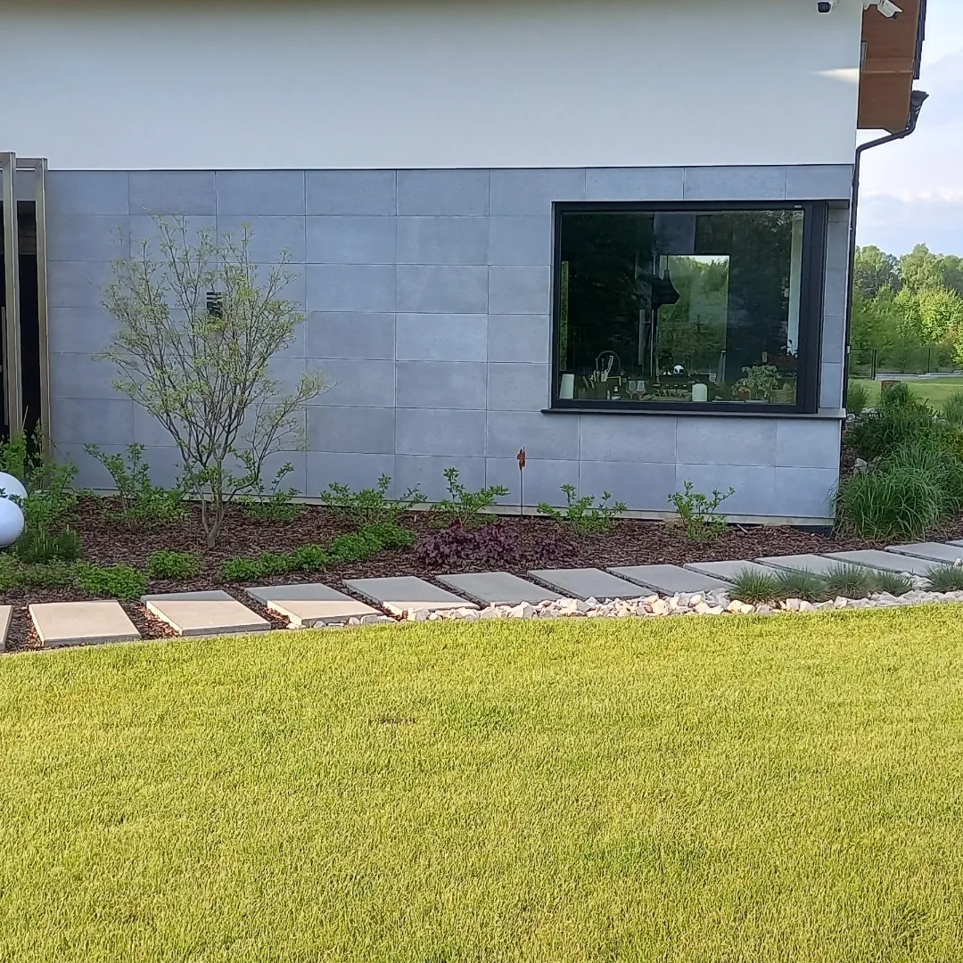 Nowoczesny ogród przydomowy z równo przystrzyżonym trawnikiem, ścieżką z płyt betonowych i rabatą z krzewami pod oknem domu wykończonego szarą płytką.