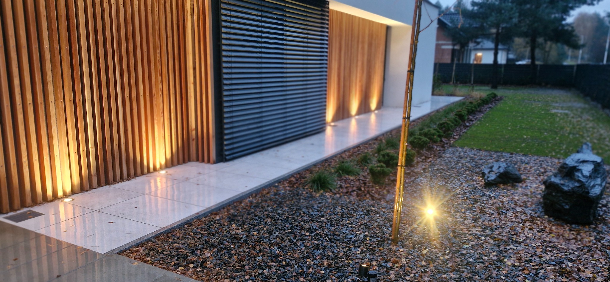 Nowoczesny ogród z podświetloną elewacją drewnianą i kamienną rabatą. Minimalistyczny design z elementami oświetlenia punktowego i dekoracyjnymi kamieniami.