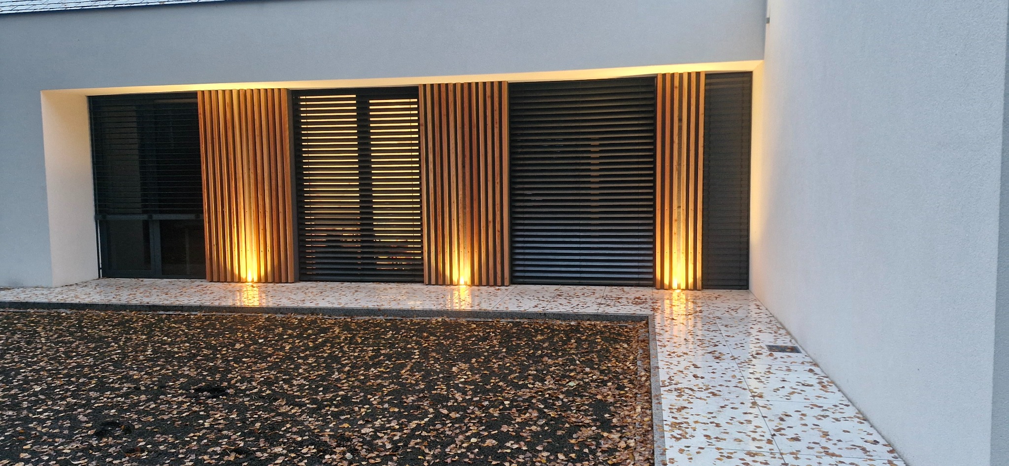 Nowoczesna fasada budynku z drewnianymi panelami i oświetleniem punktowym, liście na ziemi. Minimalistyczny design z akcentami naturalnego drewna.