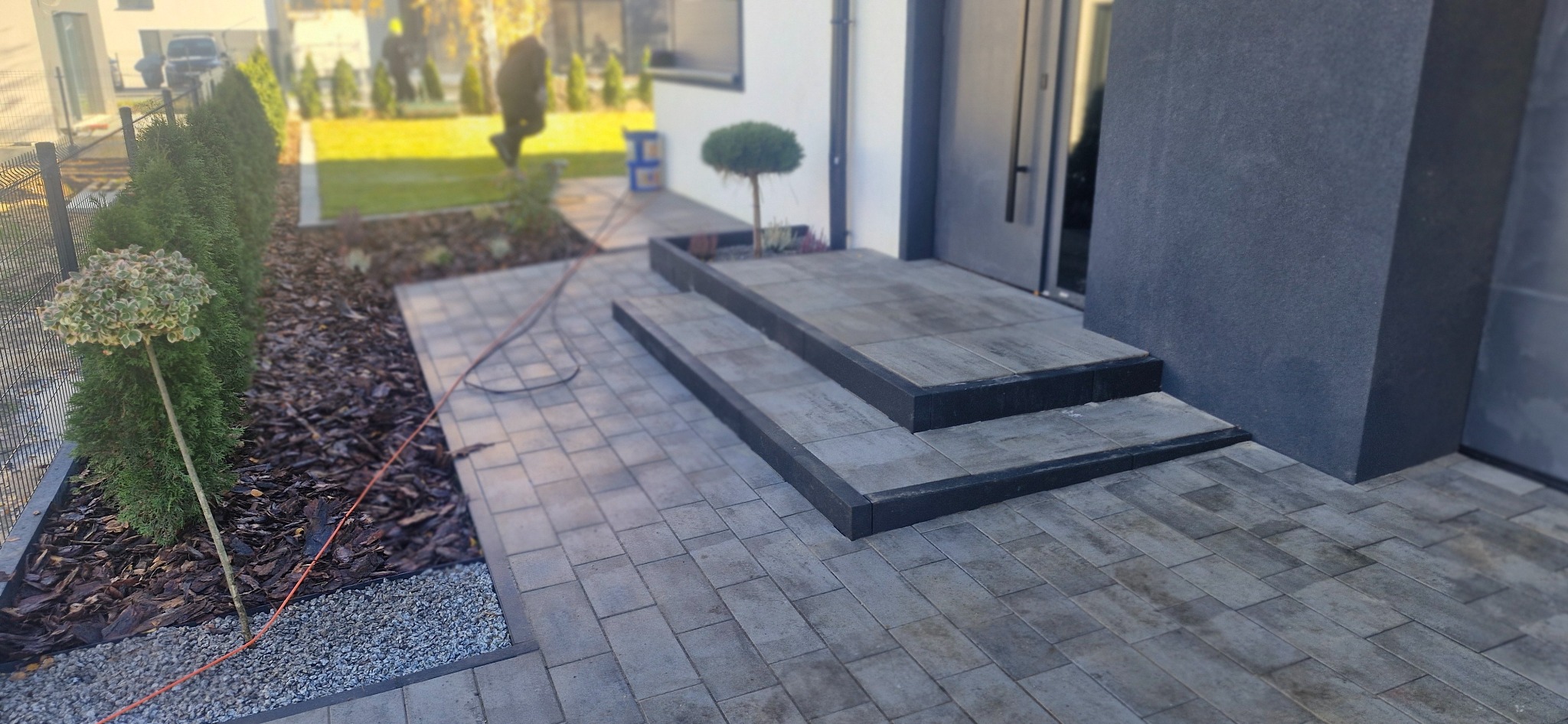 Nowoczesny ogród z szarą kostką brukową prowadzącą do wejścia, obrzeża z kory i kamienia, oraz rzędem tuj wzdłuż ogrodzenia. Minimalistyczny design.