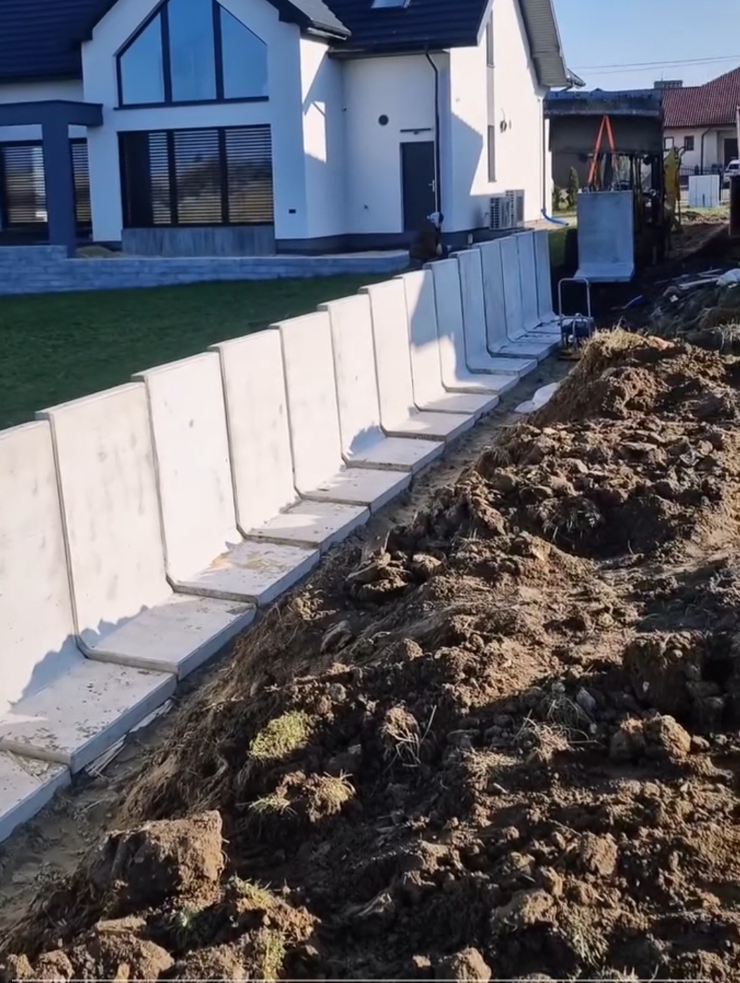 Montaż prefabrykowanych elementów betonowych na skarpie przy nowoczesnym domu. Widoczna ziemia i sprzęt budowlany. Etap prac ziemnych i wzmacniania terenu.