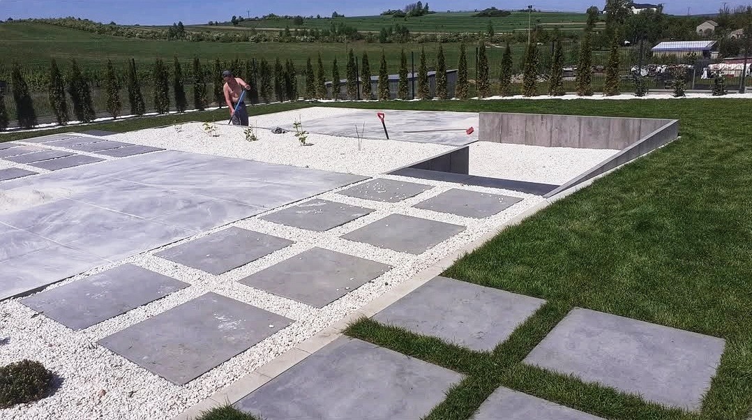 Nowoczesny ogród w trakcie realizacji: szare płyty chodnikowe ułożone w trawie, biały żwir, betonowe obrzeża, rząd tuj i pracownik z grabiami w tle.