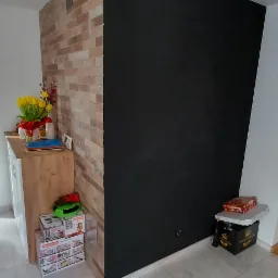 Wnętrze domu w Rząśni z wykończoną ścianą z cegiełek, obok której stoi komoda z kwiatami i dekoracjami, oraz czarna ściana z niewielkim stolikiem i pudełkami, a w tle widoczny pies rasy Mops.