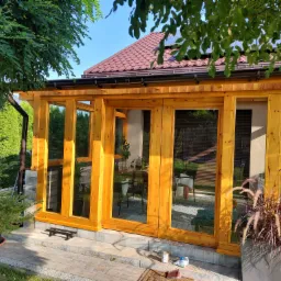 Drewniana konstrukcja z dużymi oknami, prawdopodobnie altana lub pergola, z widocznym wyposażeniem wnętrza i roślinami ozdobnymi na zewnątrz, w słoneczny dzień.