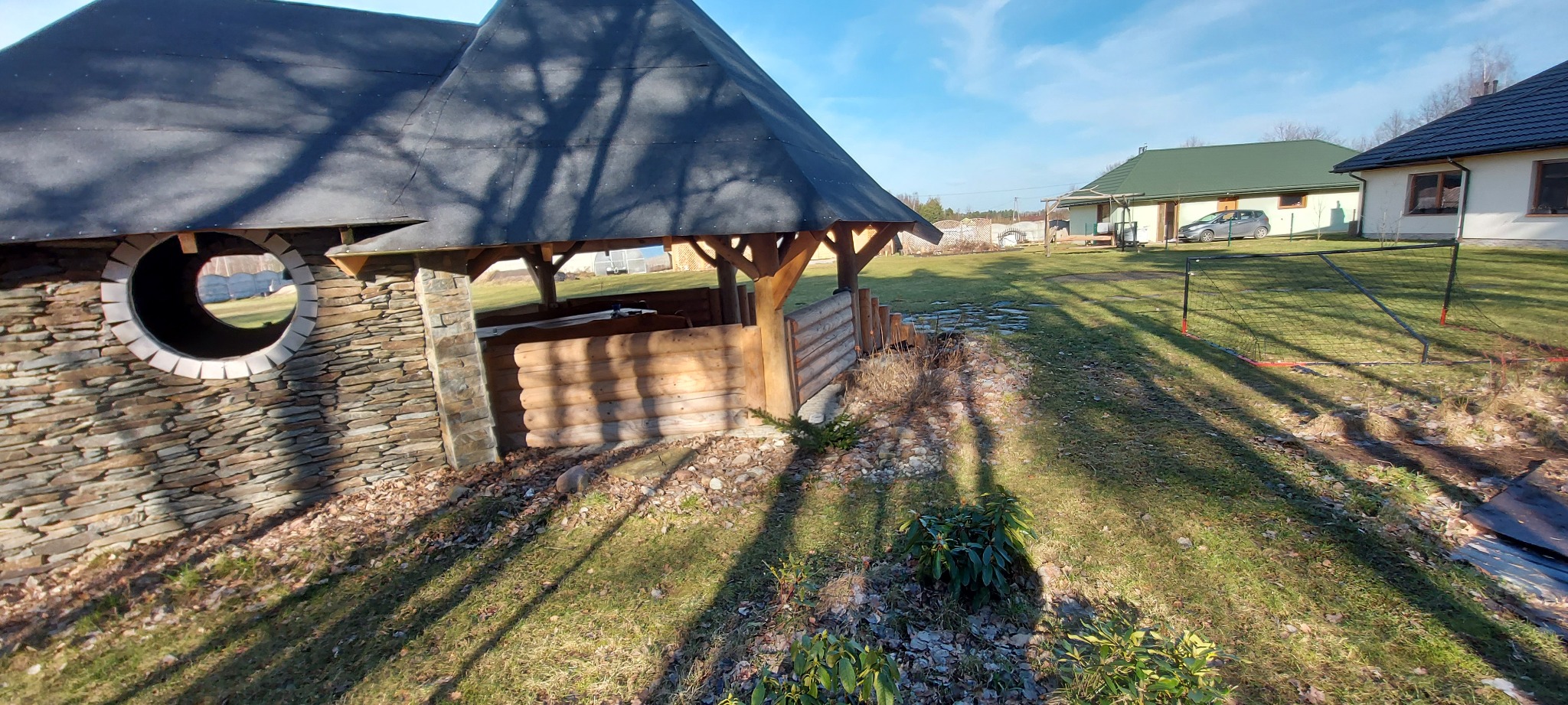 Altana z kamienną ścianą z okrągłym oknem i drewnianą konstrukcją, widok na trawnik i dom w tle.