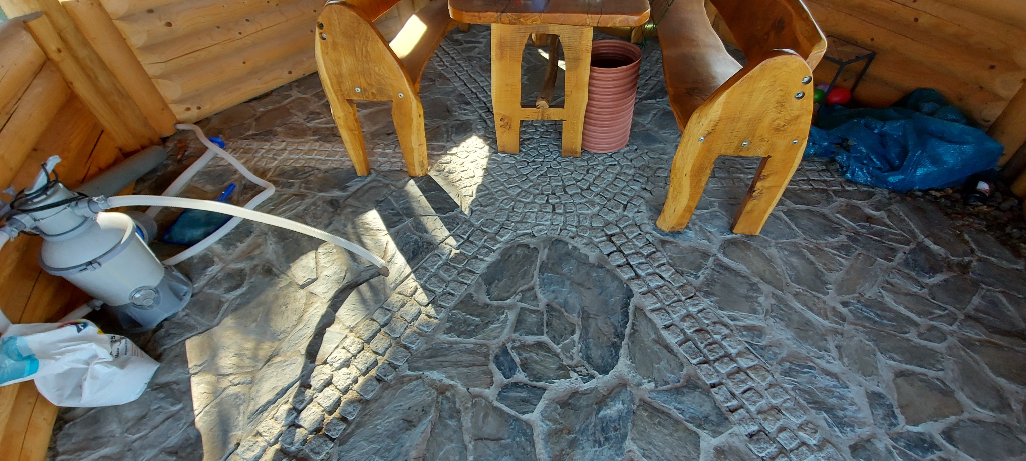 Wnętrze drewnianej konstrukcji z kamienną podłogą, na której stoi drewniany stół i krzesła, widoczny filtr do basenu i niebieska folia. Podłoga wyłożona kamieniami o różnych kształtach i fakturach.