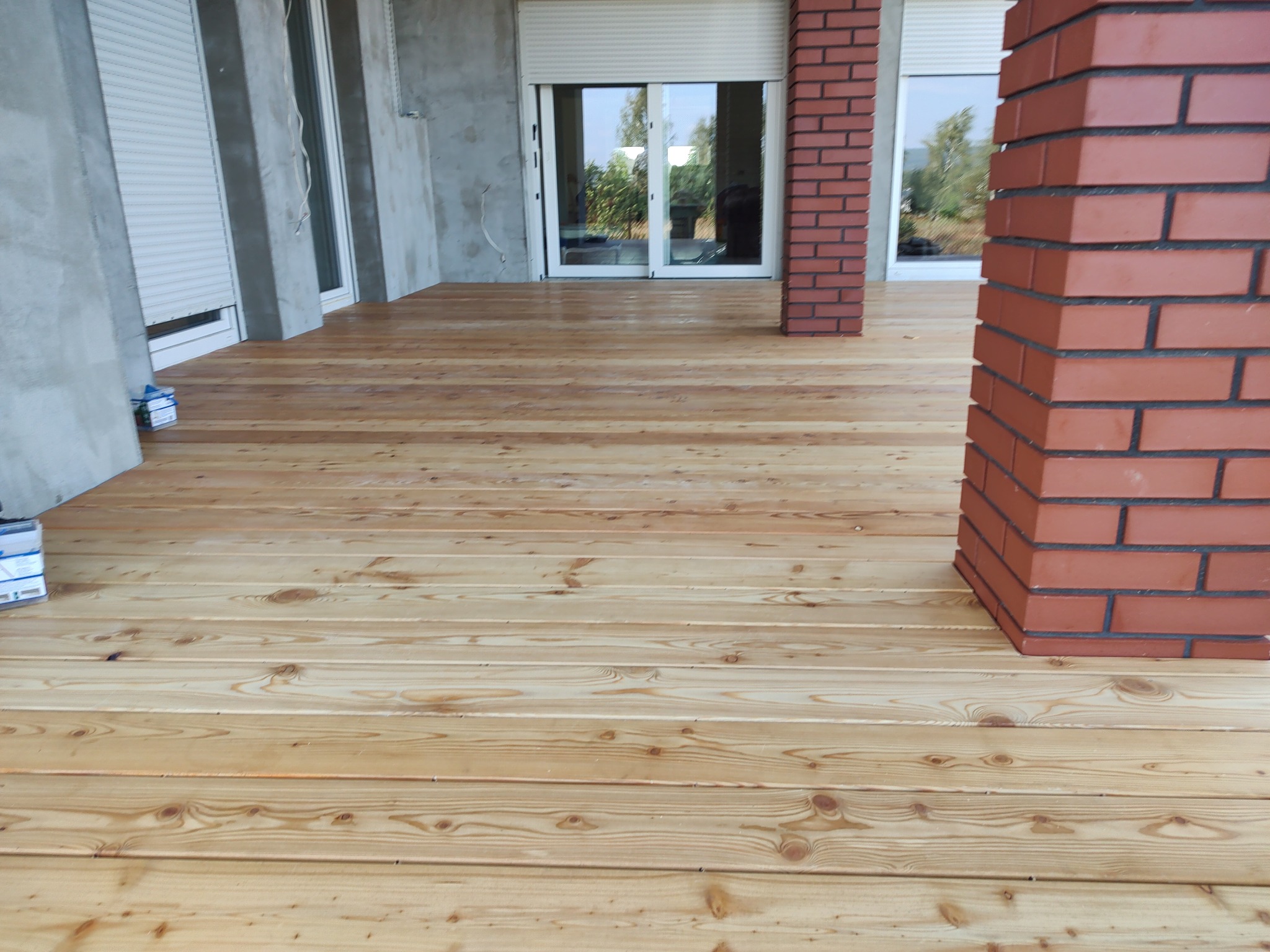 Nowy taras z jasnego drewna z widocznymi słojami, częściowo osłonięty, z ceglanymi filarami i podwójnymi drzwiami przesuwnymi w tle.