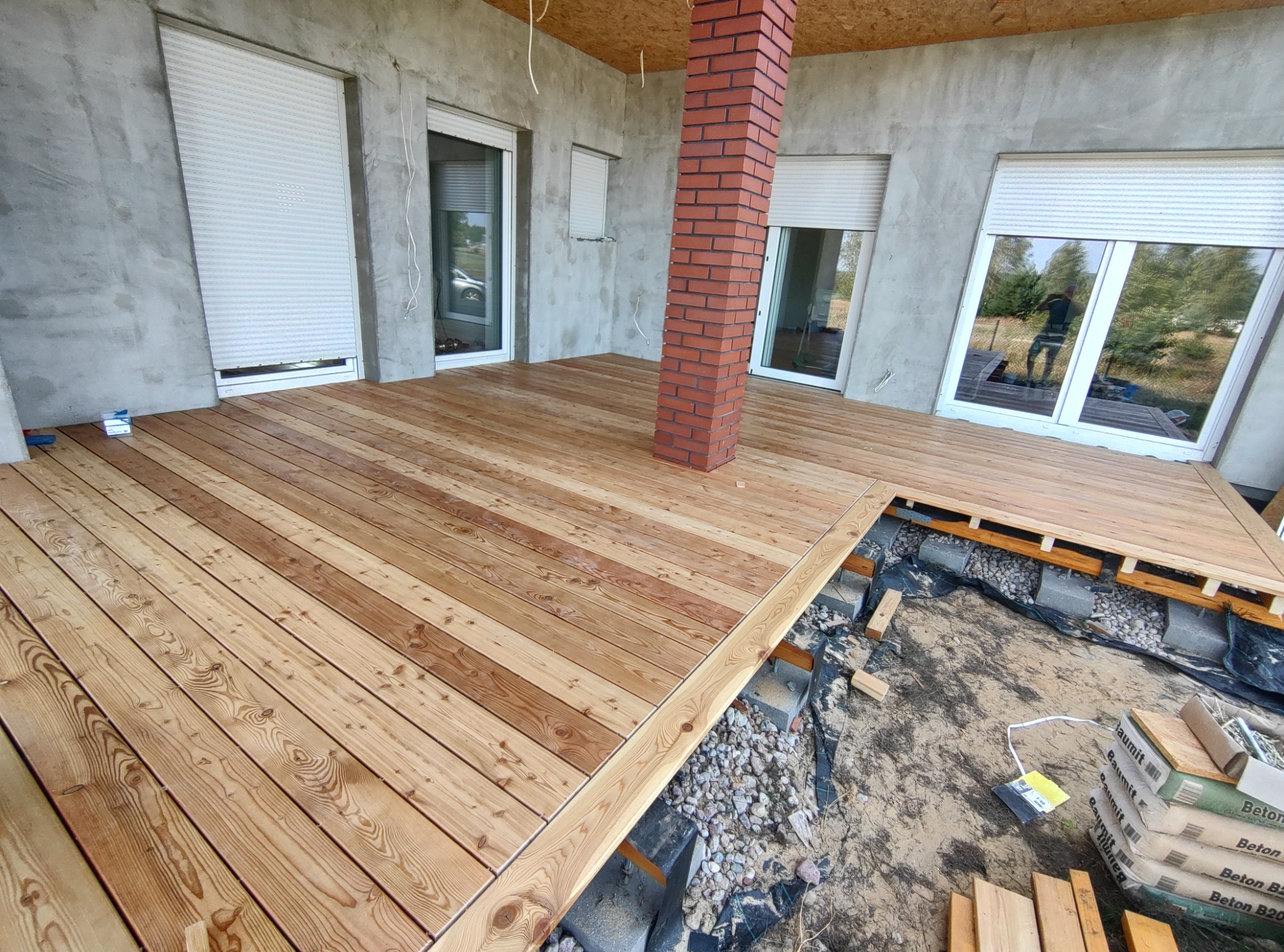 Częściowo ukończony taras z desek z widocznym podłożem z betonu i kamieni, ceglana kolumna, szare ściany budynku z oknami i roletami.