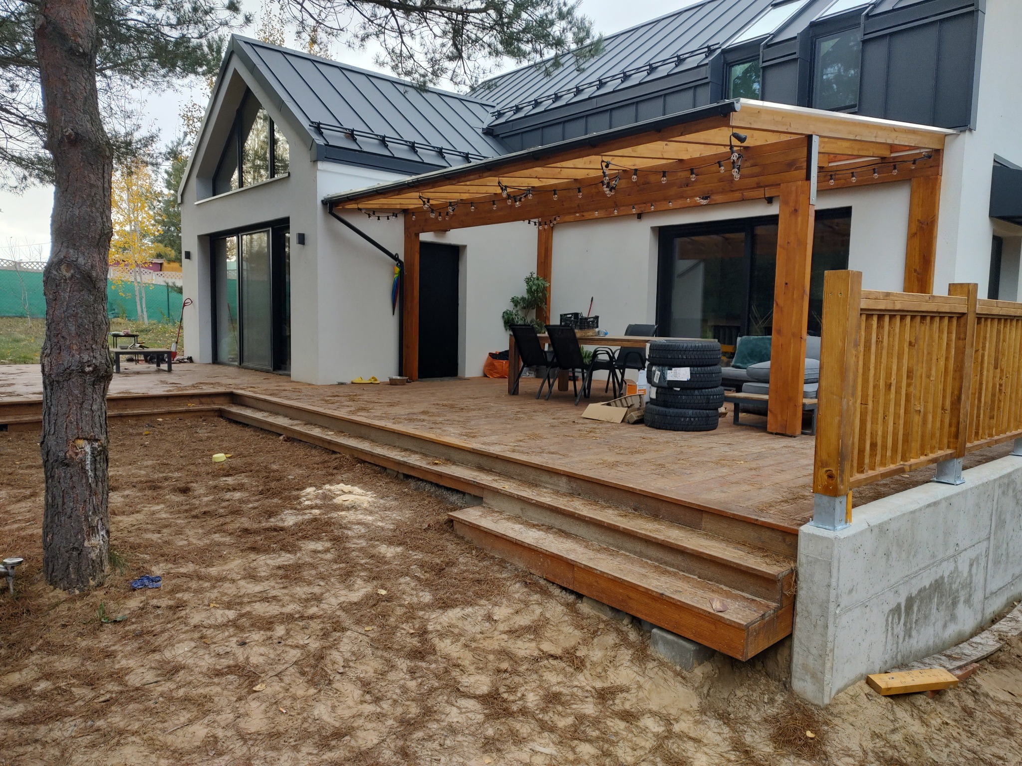 Drewniany taras z pergolą i schodami przed nowoczesnym domem z szarą blachodachówką, oświetlony girlandami żarówek, z widocznymi śladami prac ziemnych wokół.