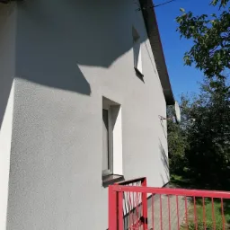 Jasnoszara elewacja domu jednorodzinnego z oknami i fragmentem czerwonego ogrodzenia, widok z boku w słoneczny dzień.