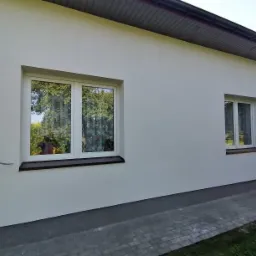 Świeżo otynkowana elewacja budynku jednorodzinnego w kolorze białym, z widocznymi dwoma oknami z brązowymi parapetami i szarym cokołem, kostka brukowa wzdłuż ściany.